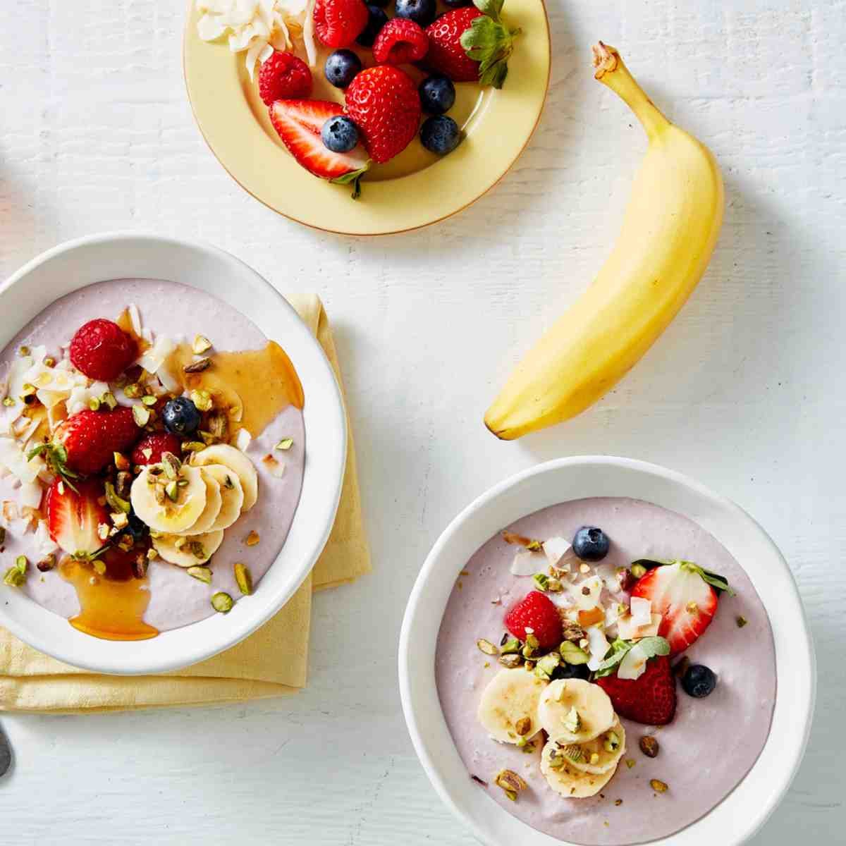 Two bowls filled with a dairy-free smoothie topped with fruits and bananas. On the top is a Havana banana, and a plate filled with cut berries.