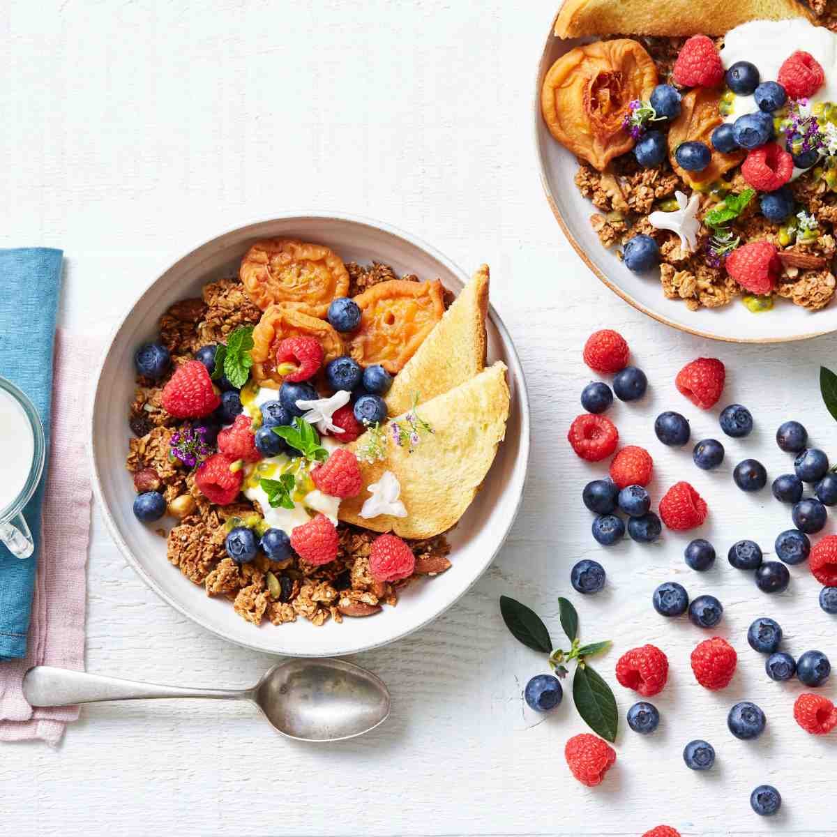Two bowls of granola topped with berries and edible flowers with brioche bread on the side of the bowl.