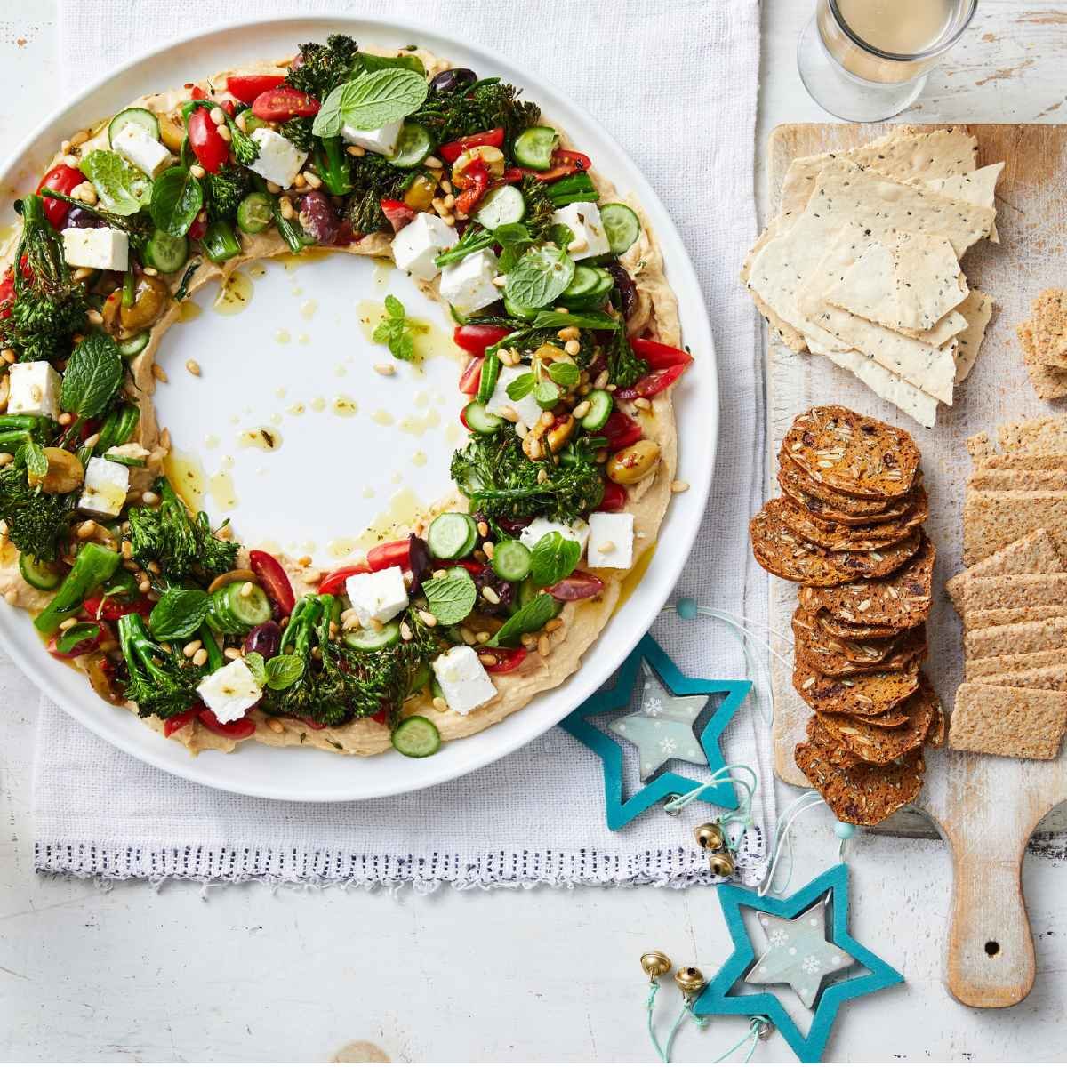 A holiday wreath on a white plate made out of hummus, topped with roasted broccolini,feta, olives and pine nuts.