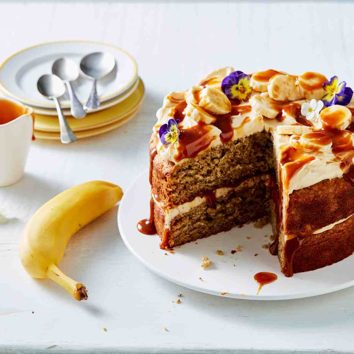 Layered banana cake topped with cream, dulce de leche, and flowers on a white plate with a Havana banana next to it.