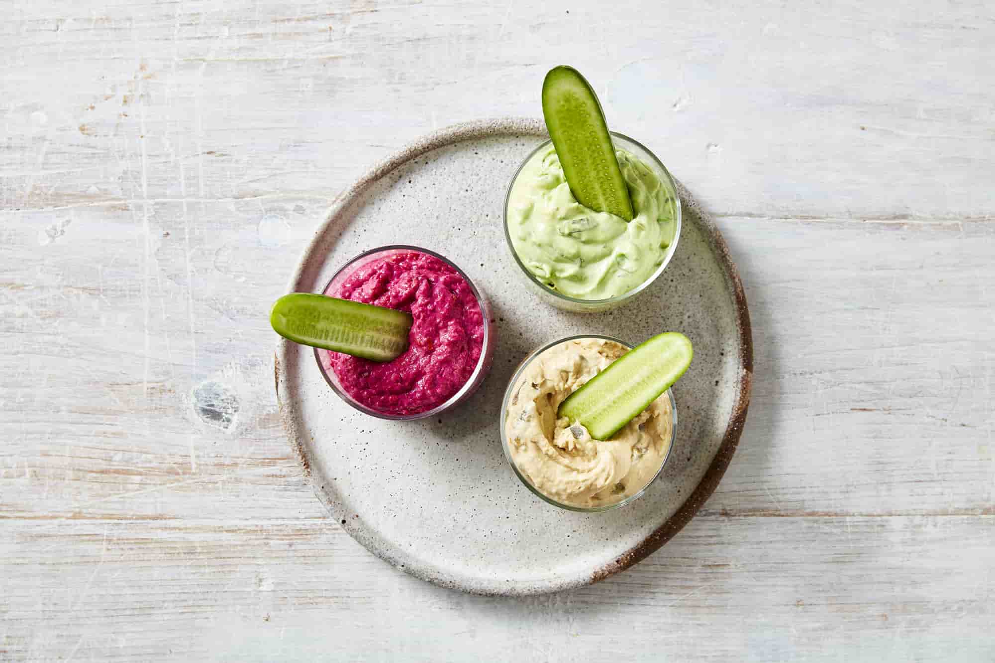 Three types of dips in mini bowls with sliced cucumbers on a white plate.