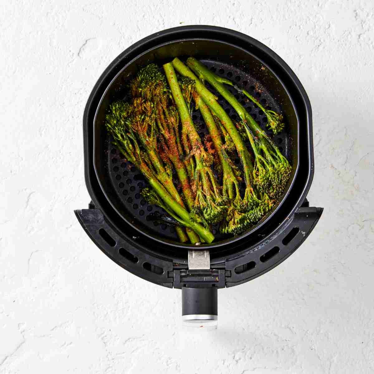 Raw broccolini in an air fryer basked sprinkled with all purpose seasoning.