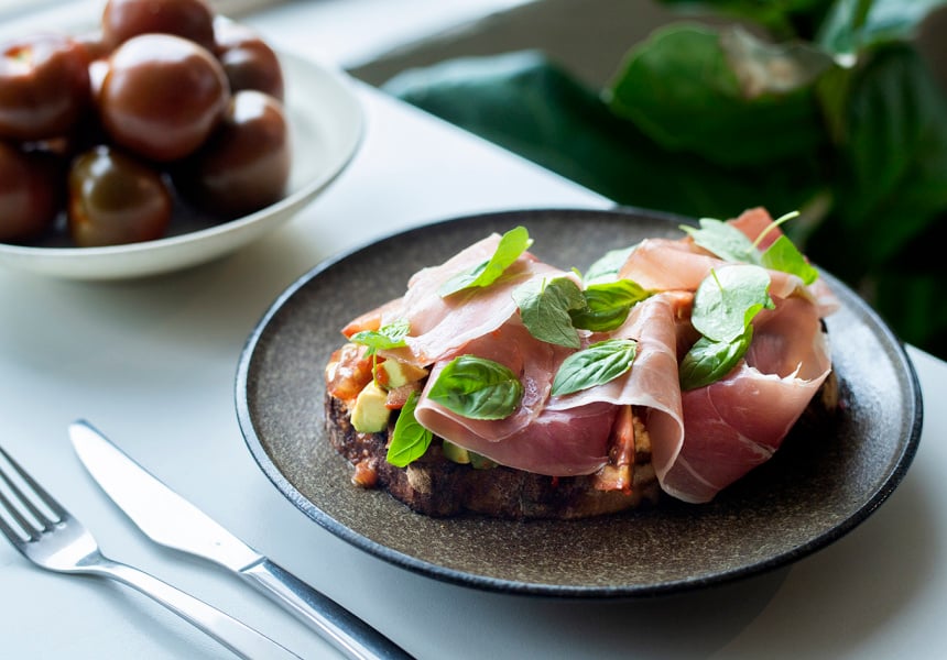 A slate gray plate with a slice of bread topped with tomatoes and prosciutto. 
