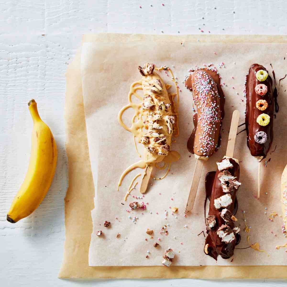 Little Gem banana pops coated with chocolate and candy on baking paper, with a banana next to them.