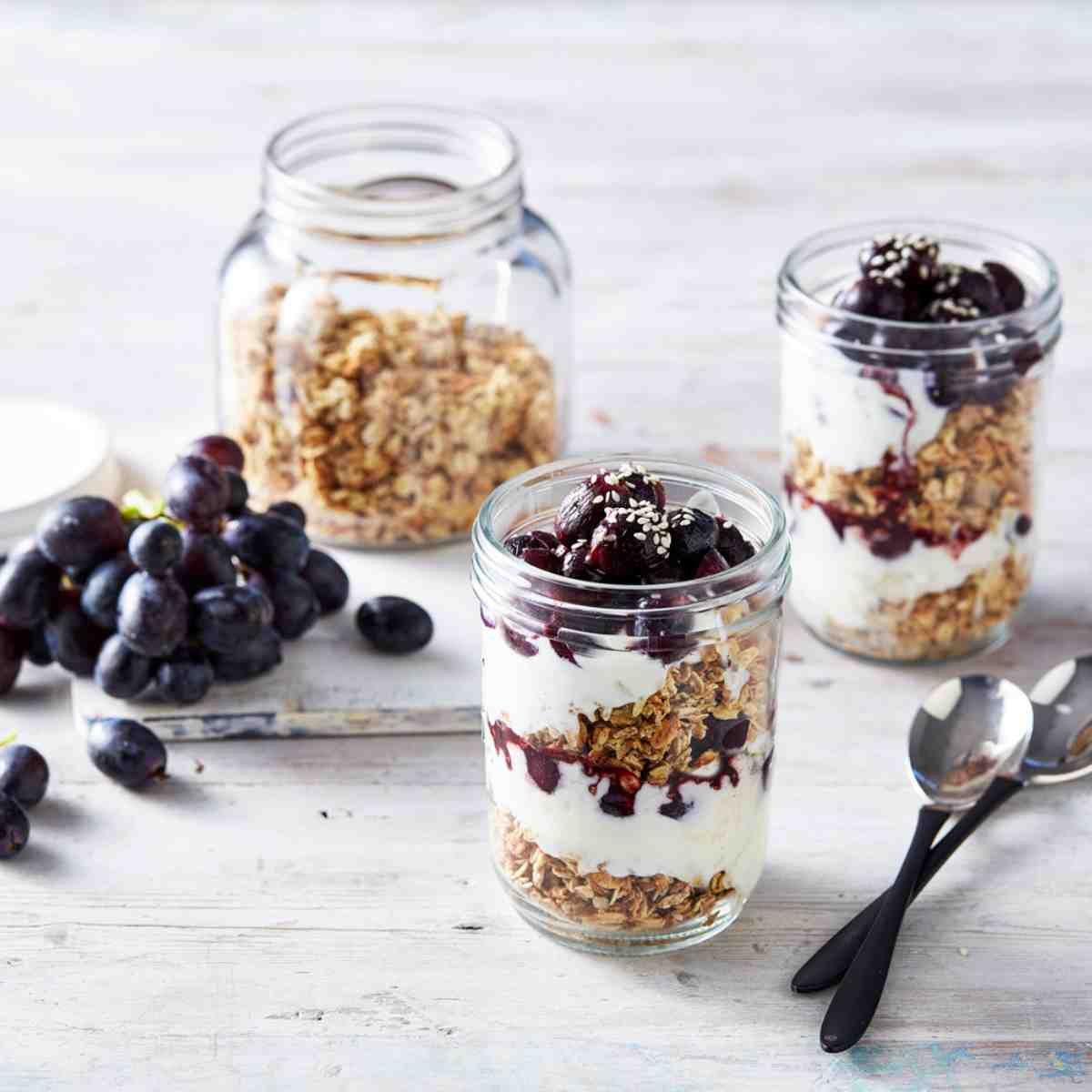 Two jars layered with granola, yoghurt and roasted grape compote with a jar of granola and a bunch of grapes in the background.