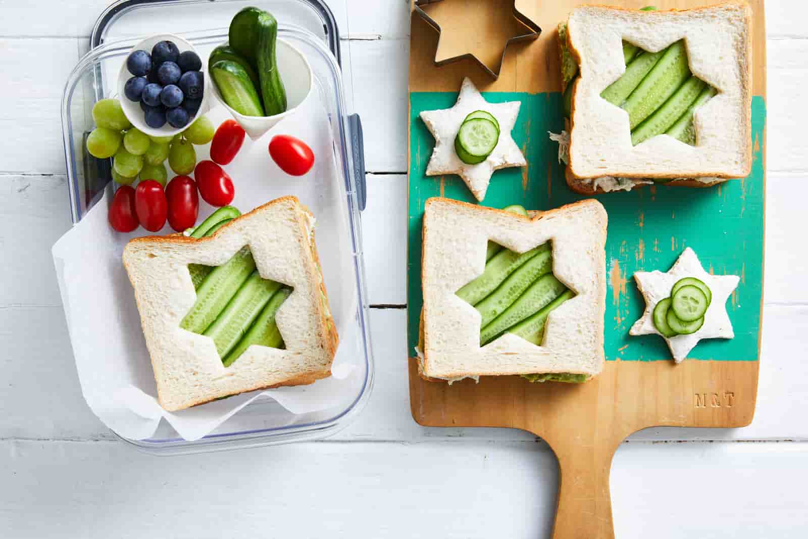 Star shaped sandwiches along with fruits and vegetables in a lunchbox. Next to it is a board with another sandwich and a star shaped cutter.