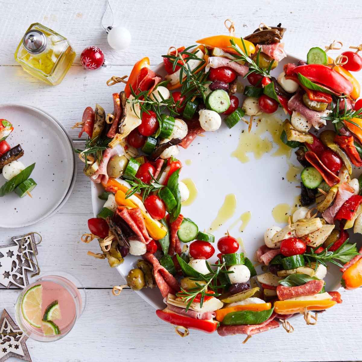 A wreath made out of antipasto, charcuterie, solanato tomatoes, and qukes cucuumebrs on a white plate. 