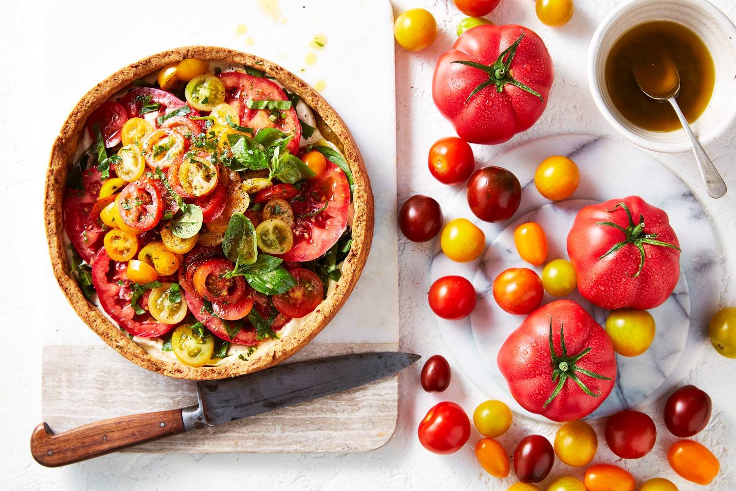 Pastry tart with sliced tomatoes, and basil with a knife next to it. On the right are a variety of tomatoes.