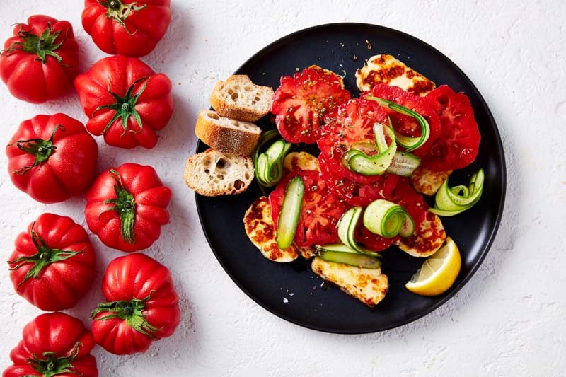 Haloumi topped with monterosa tomatoes on a black plate, with more tomatoes and crispbread on the side.
