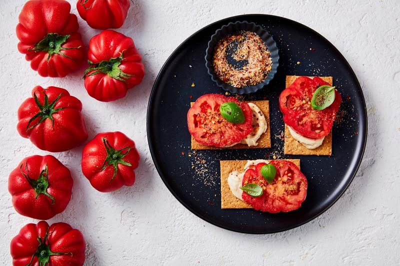 Crispbread with sliced monterosa tomatoes on a black plate, and more tomatoes on the side.