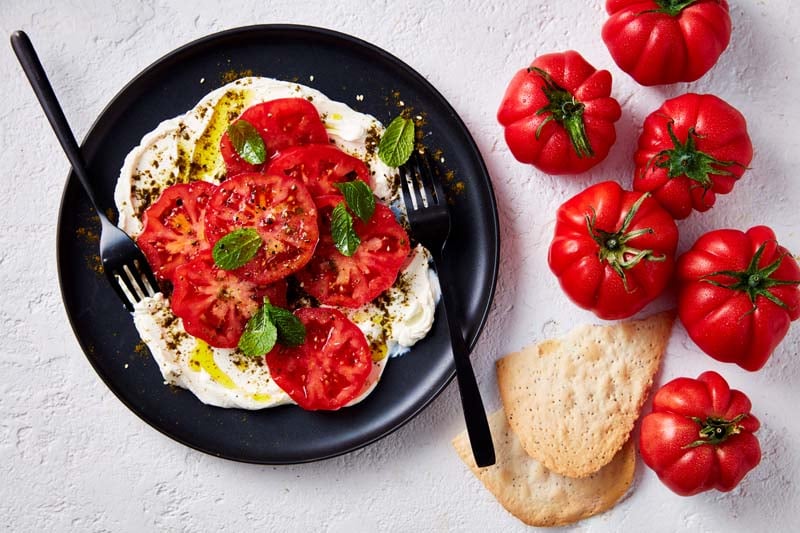 Labneh topped with monterosa tomatoes on a black plate, with more tomatoes and crispbread on the side.