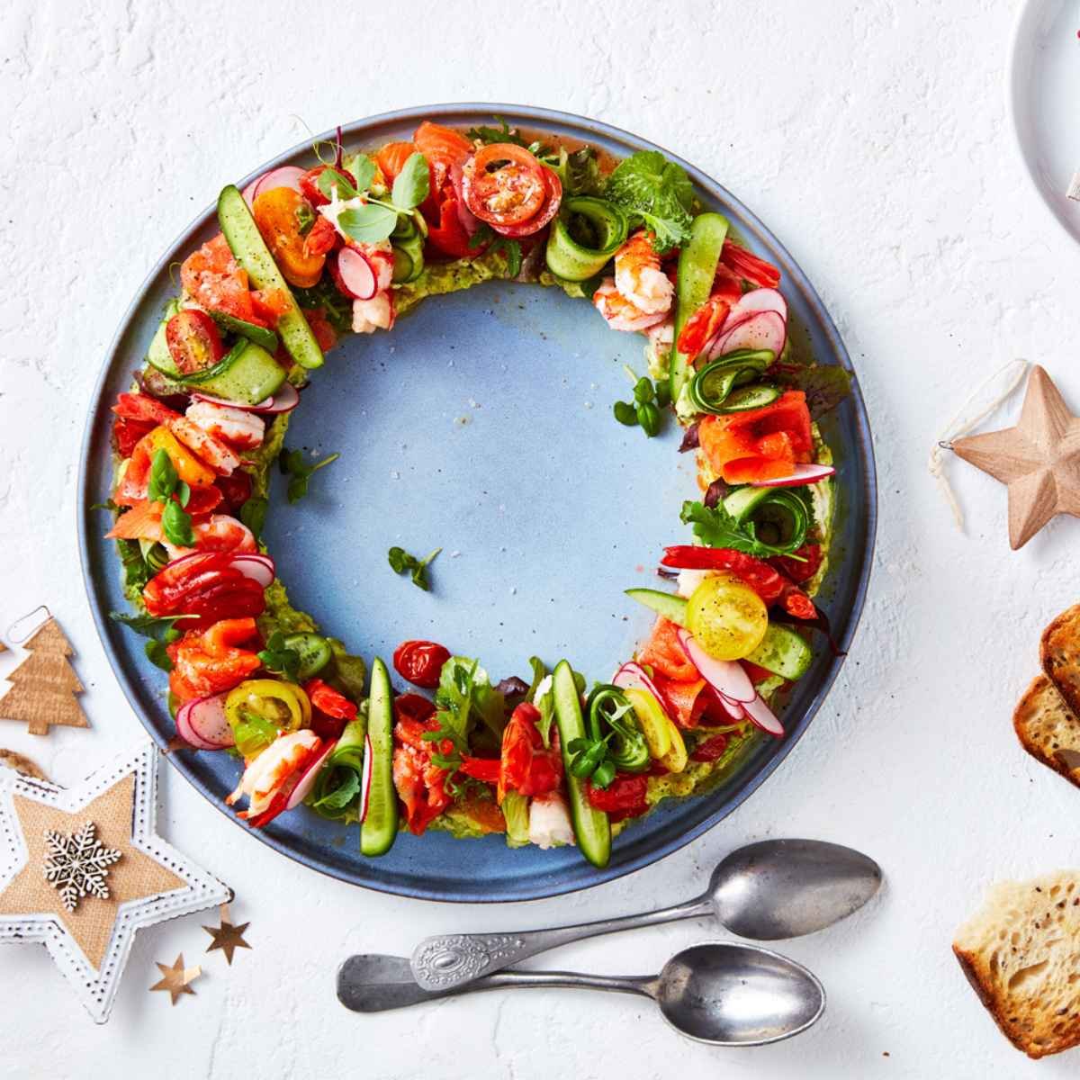 A wreath made with prawns, salmon, Qukes cucumbers, mix-a-mato tomatoes and petite tomatoes. On the side are spoons and Christmas ornaments.