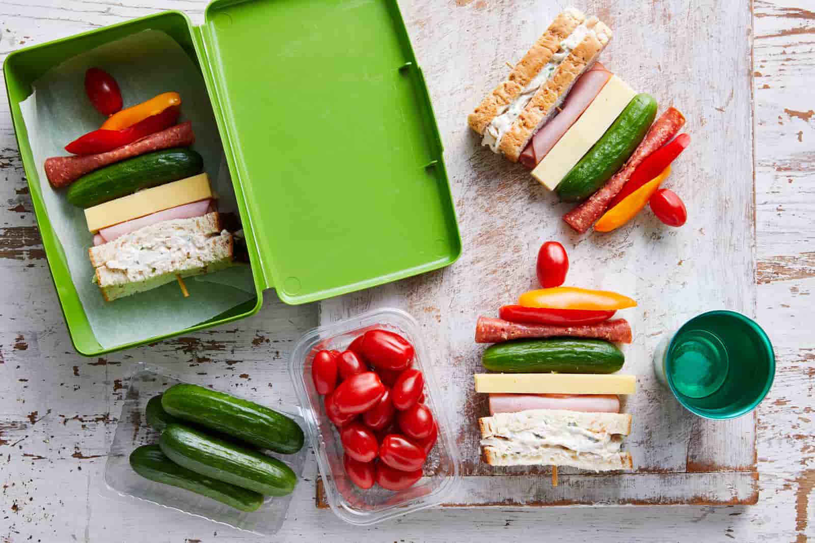 Sandwiches on a skewer in a green lunchbox and outside, with baby cucumbers and snacking tomatoes next to them.
