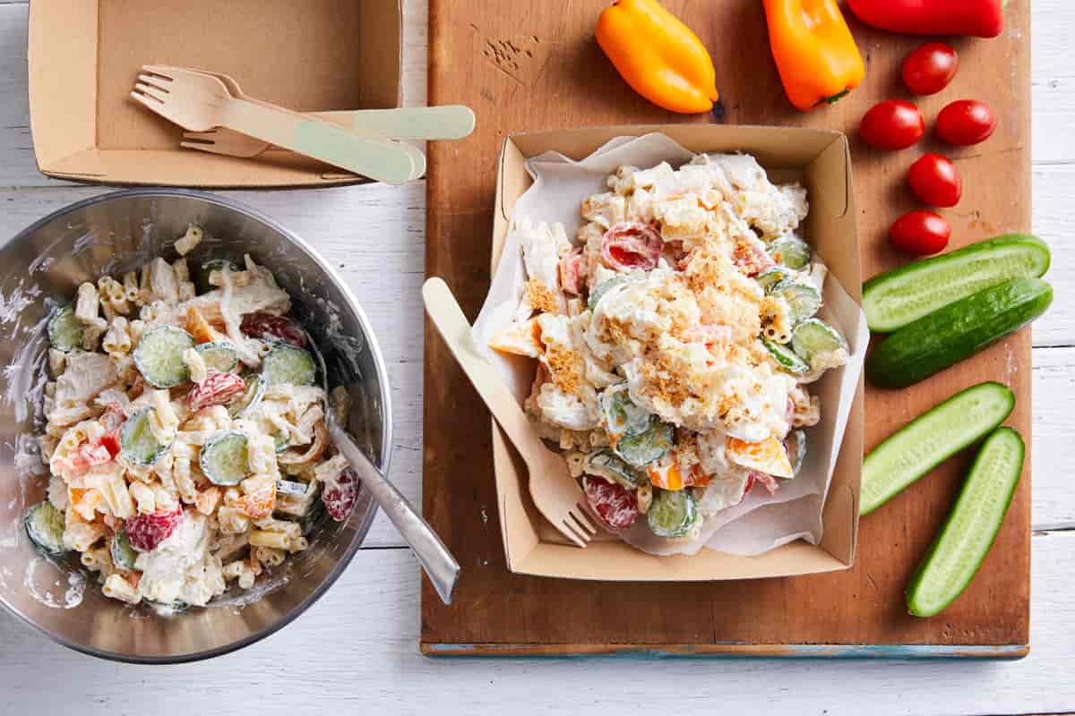 Mac N Cheese salad in a cardboard box, with a bowl and vegetables next to it.