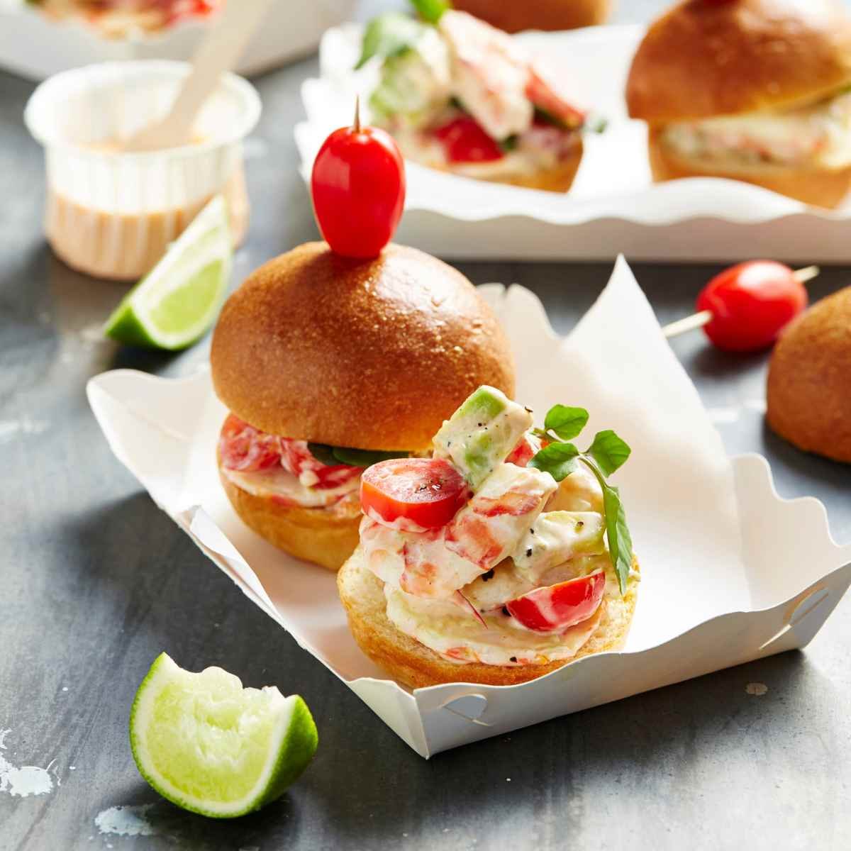 Two slider rolls filled with prawn, solanato tomatoes and avocado in a white paper container.
