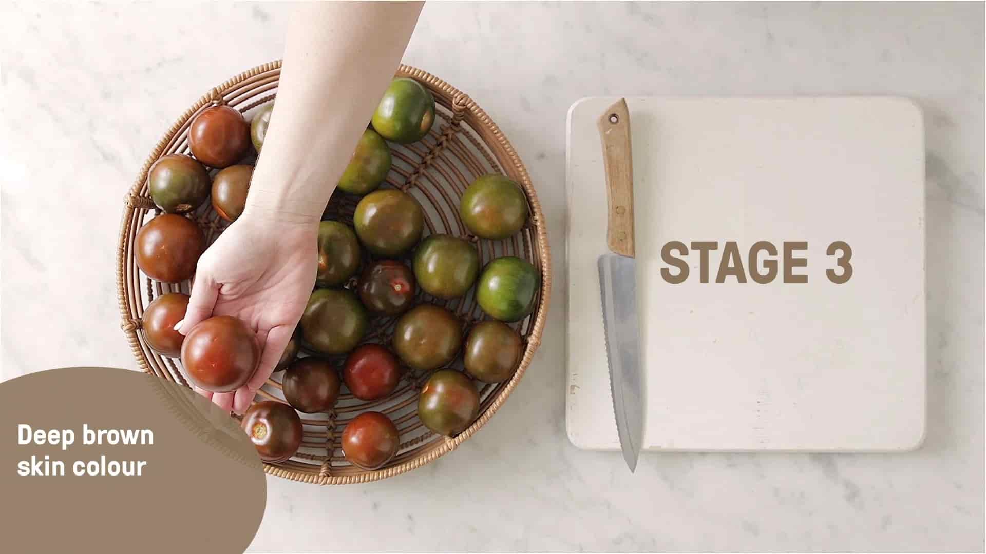 A hand holding a red kumato tomato over a basket of kumatoes.