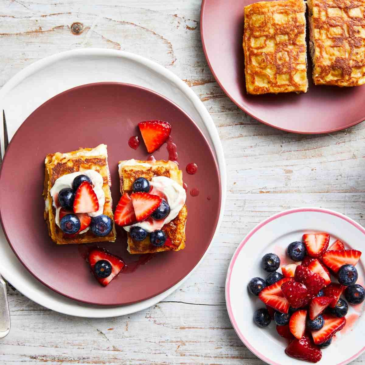 A stack of waffles topped with yoghurt and fruit on a pink plate. Next to the plate are more berries, and waffles.