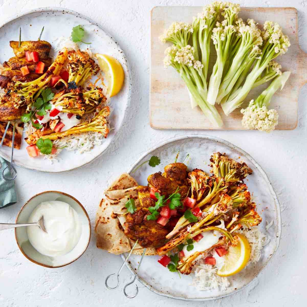 Tandoori roasted cauli-blossom with BBQ chicken skewers, lemon, veggies served with rice and roti on two white plates.