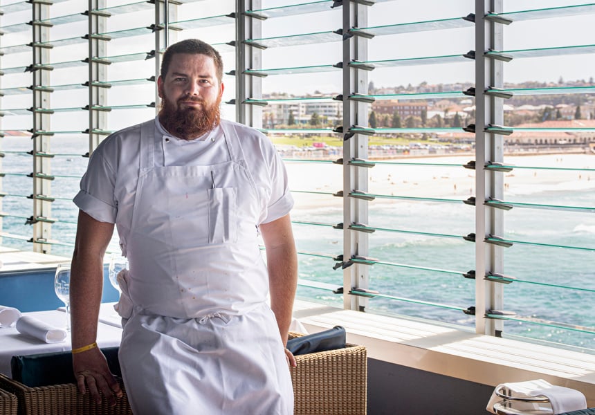 A man wearing an chef's jacket standing next to a table. 