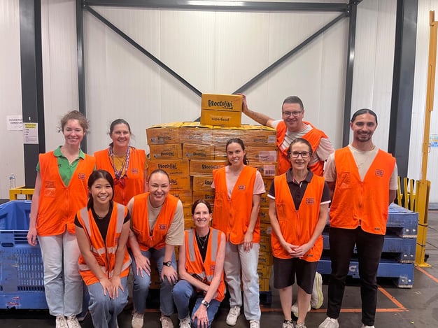 9 people of various ages and genders wearing high vis vests. In the background is stacked pallets and boxes of Perfection Fresh Broccolini