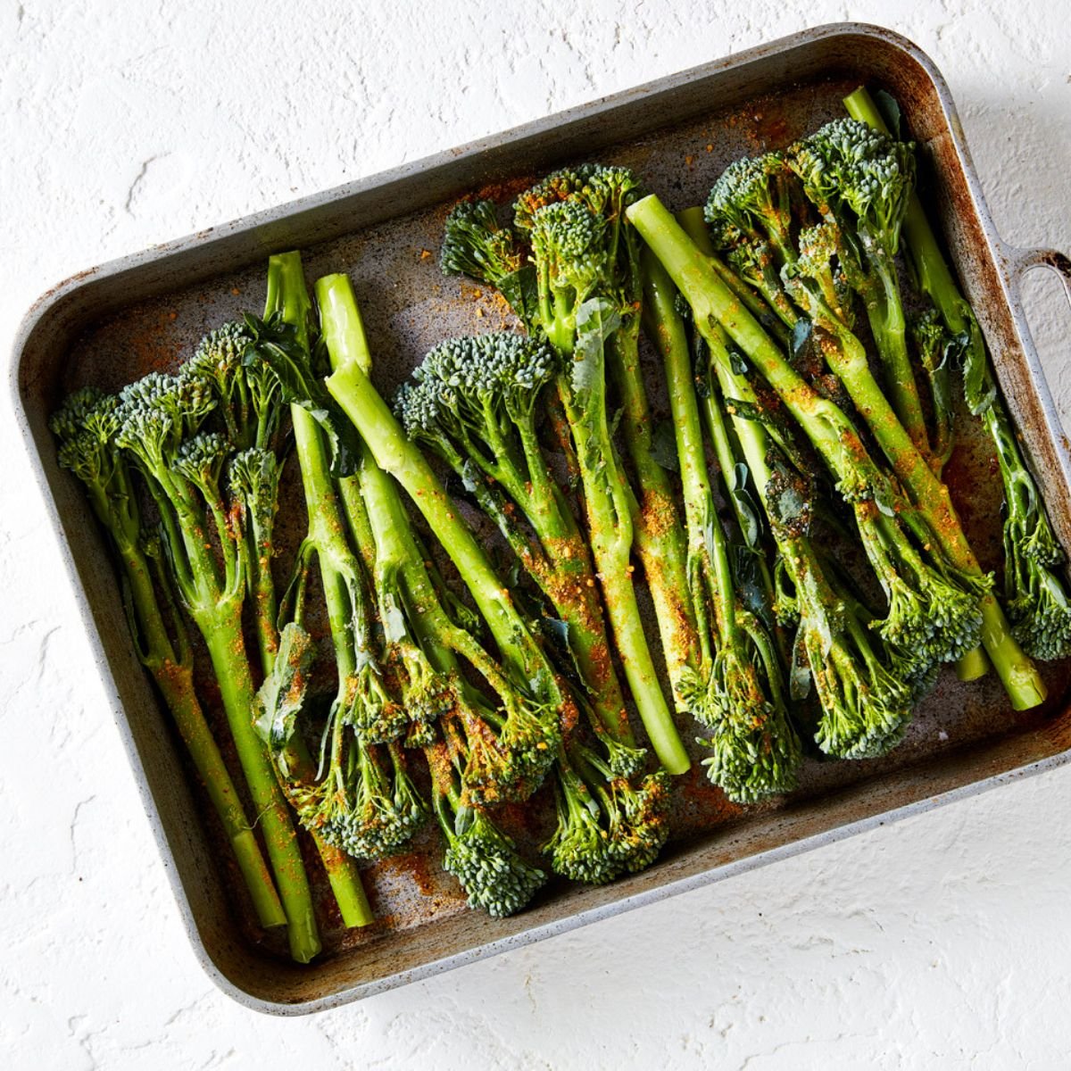 Raw broccolini placed in an oven roasting tray, and sprinkled with seasonings.