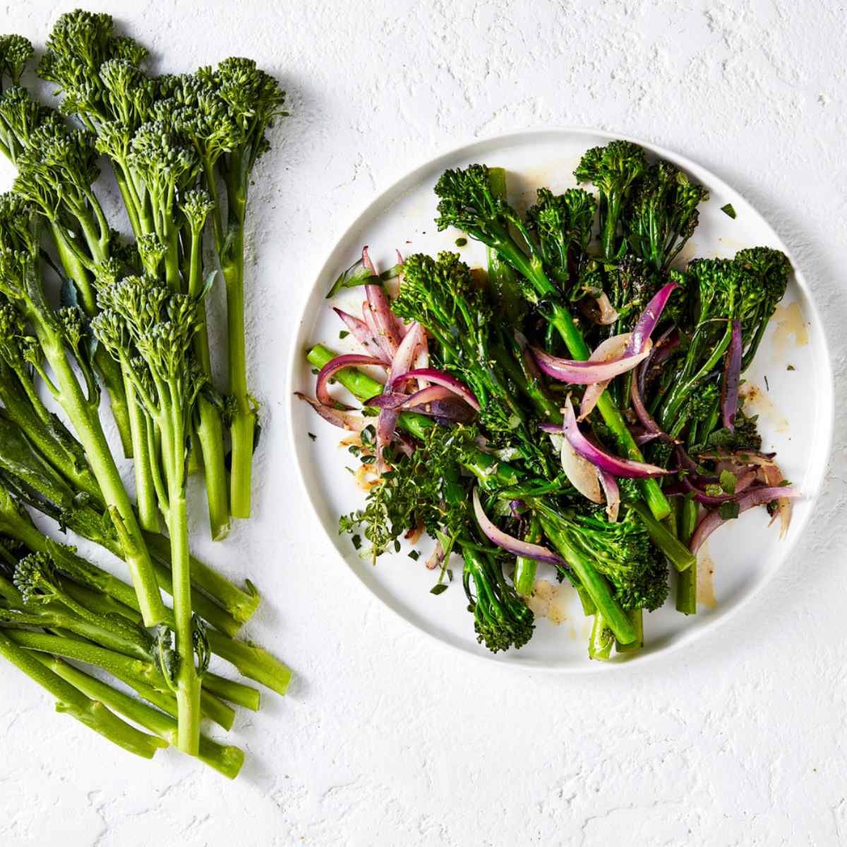 Sauteed broccolini with onions on a white plate with a bunch of Broccolini next to it. 