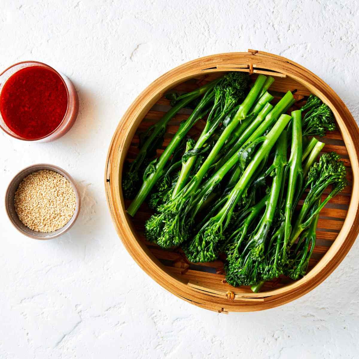 Steamed Broccolini in a wooden steamer basket with a bowl of sesame seeds, and gochujang dressing on the side.