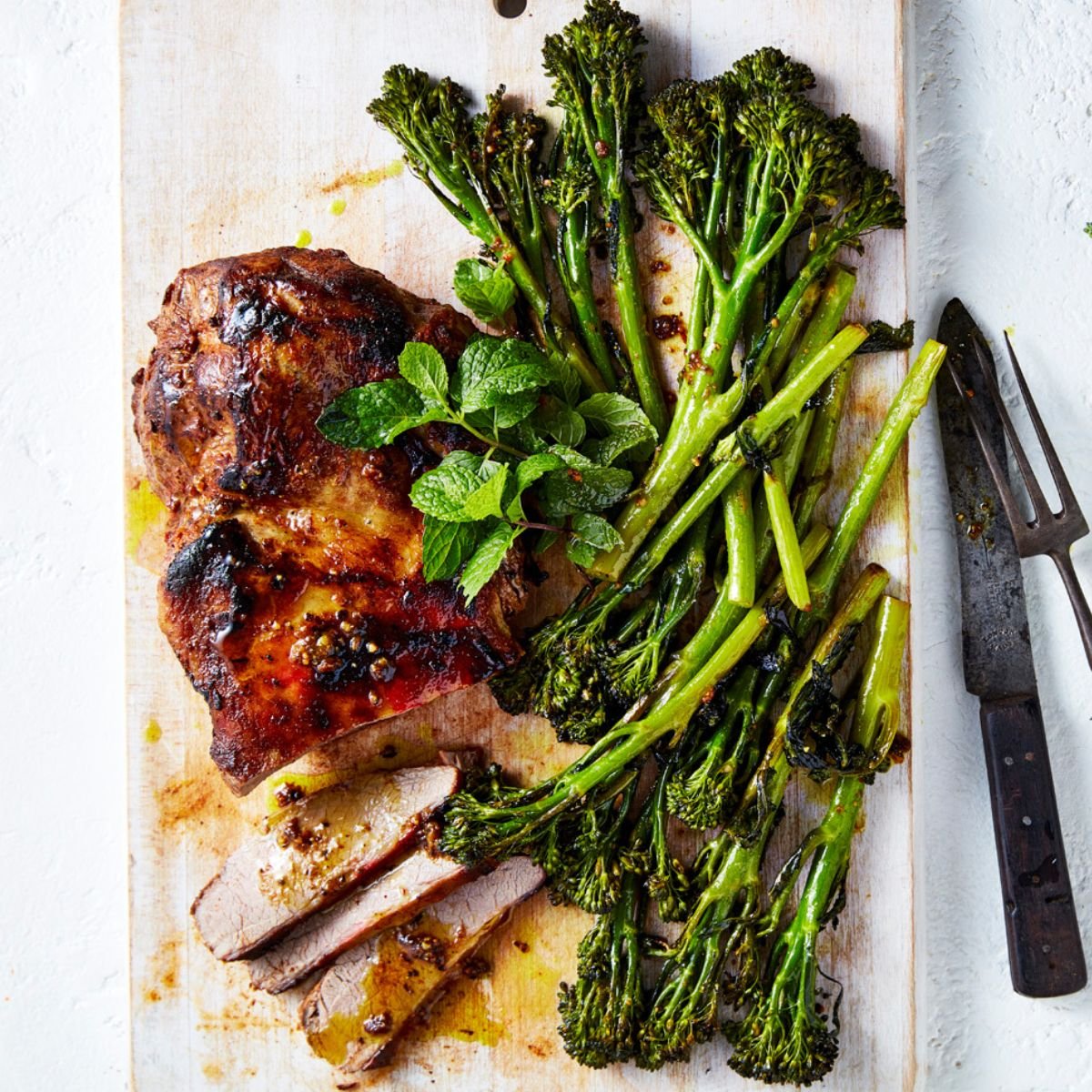 Roasted broccolini with roast lamb on a white serving board with a knife and a fork. 