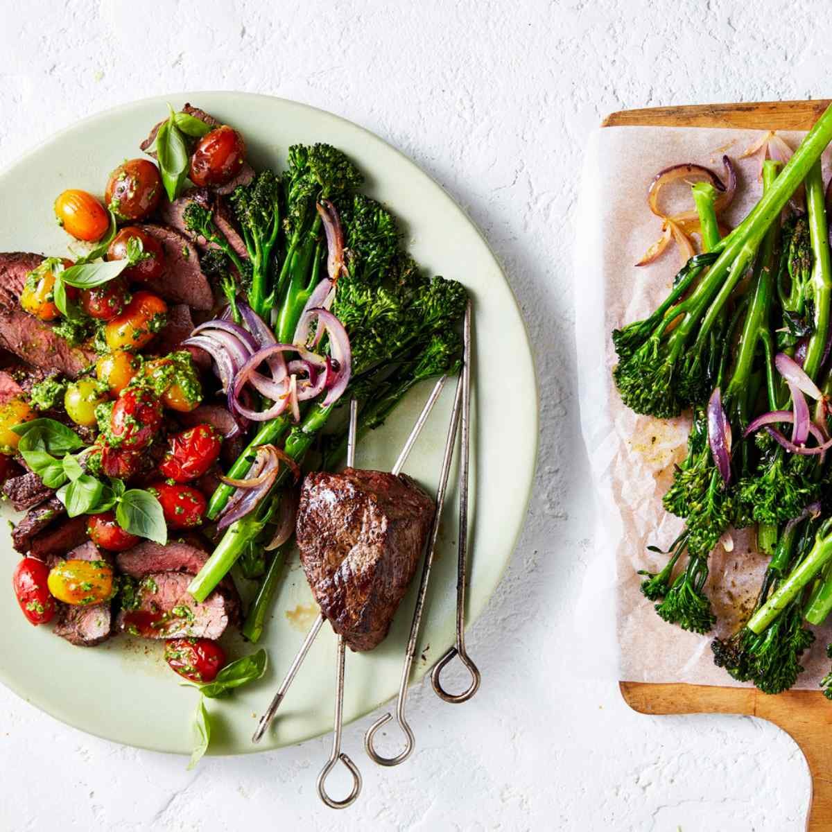 A green plate with rump roast on skewers, onions, tomatoes and Broccolini. Wooden board with Broccolini next to it.