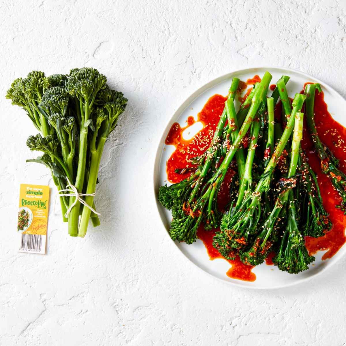 Steamed broccolini with gochujang dressing in a white plate, more loose broccolini on the left.