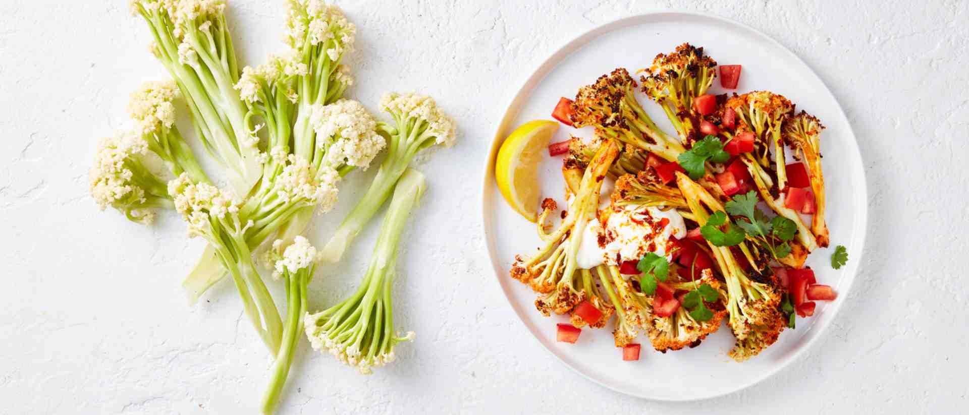 Tandoori roasted cauli-blossom on a white plate topped with yoghurt, tomatoes and lemon. On the side is raw cauli-blossom. 